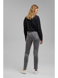 Camisa preta de mangas longas com um corte descontraído, combinada com jeans cinzentos justos. Os jeans apresentam uma textura desgastada e bolsos traseiros, complementados por ténis brancos.
