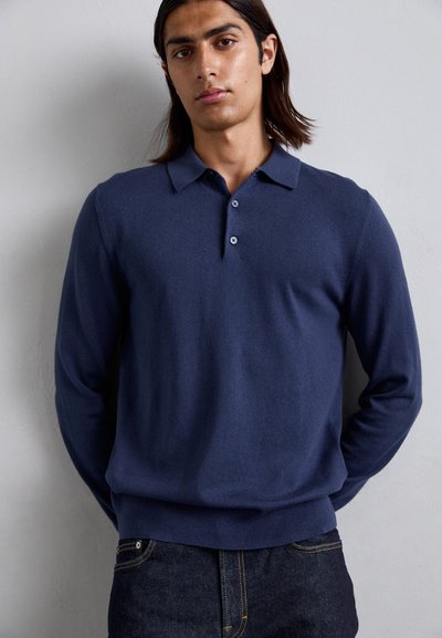 Jeune homme avec des cheveux longs jusqu'aux épaules portant un polo à manches longues bleu marine et un jean foncé, se tenant contre un mur clair.