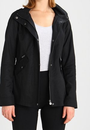 Kvinde iført en sort lynlåstrøje med klaplommer, over en lysegrå top og sorte bukser, stående mod en ensfarvet baggrund.