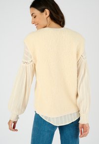 Suéter de punto texturizado crema, de corte ancho y con mangas cortadas. Superpuesto sobre una blusa ligera con pliegues y mangas abullonadas a rayas.