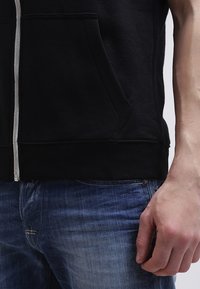 La sudadera negra con cremallera presenta una tela texturizada y un bolsillo frontal. Se lleva sobre unos vaqueros de denim azul con un acabado desgastado y detalles de costura.