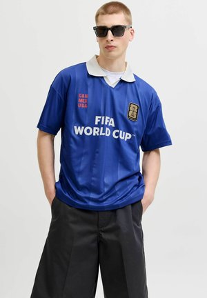 Jeune homme portant des lunettes de soleil noires et un maillot bleu de la Coupe du Monde FIFA avec l'inscription "CAN MEX USA", debout les mains dans les poches.