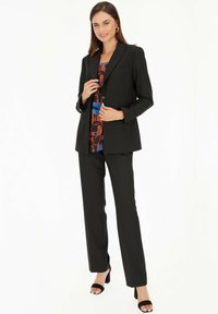 Czarny dopasowany garnitur z elegancką marynarką zapinaną na jeden guzik oraz prostymi spodniami, nałożony na wzorzystą bluzkę w niebieskie, brązowe i czerwone akcenty.