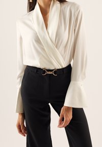 Weiße Wickelbluse mit langen, glockenförmigen Ärmeln, kombiniert mit schwarzen Hosen, die eine goldene, mondförmige Gürtelschnalle zeigen, insgesamt glatte Textur.