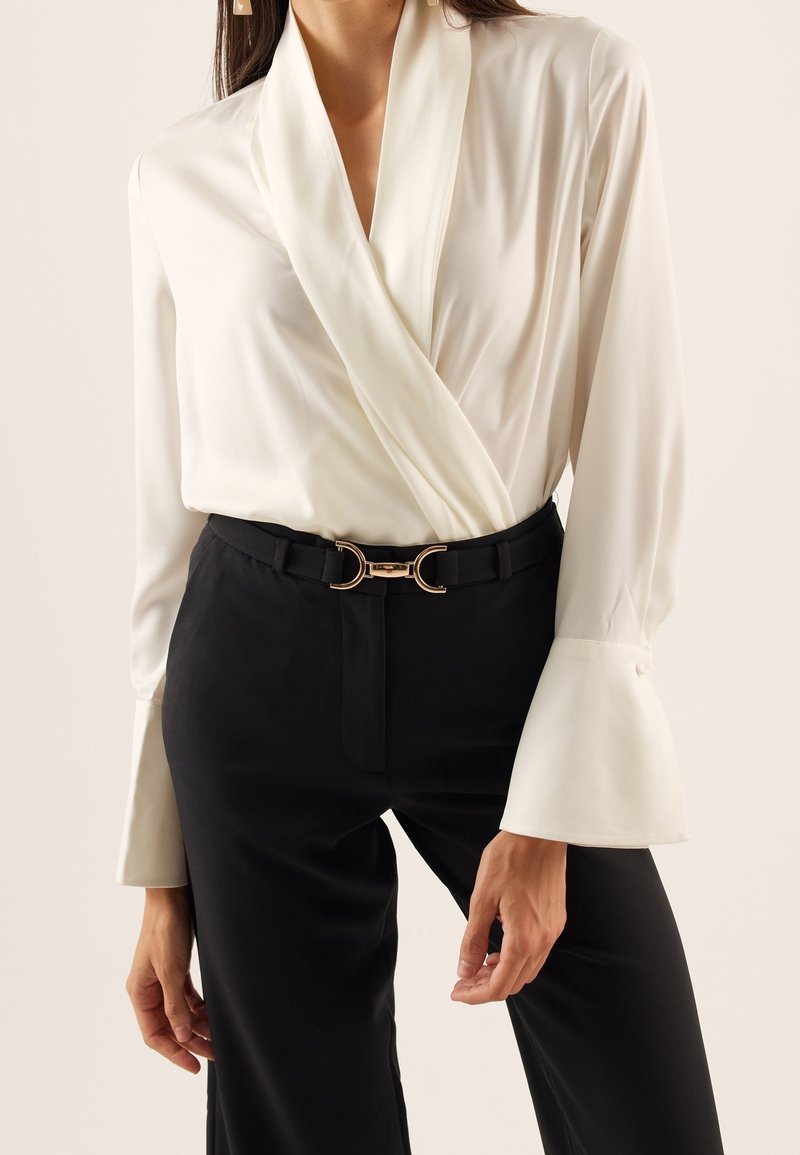 Weiße Wickelbluse mit langen, glockenförmigen Ärmeln, kombiniert mit schwarzen Hosen, die eine goldene, mondförmige Gürtelschnalle zeigen, insgesamt glatte Textur.