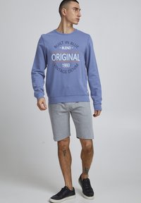 Felpa azzurra con scritta "BUILT IN BLUE ORIGINAL 1993 VINTAGE DENIM", abbinata a pantaloni corti grigi e sneaker nere.
