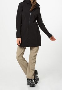 Veste imperméable noire avec capuche, fermeture éclair intégrale et deux poches latérales, associée à un pantalon beige clair et des chaussures de randonnée noires.