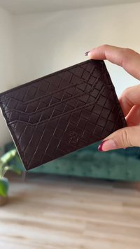 Dark brown leather card holder featuring a woven texture, slim design, and embossed logo. Displayed in hand against a light background.