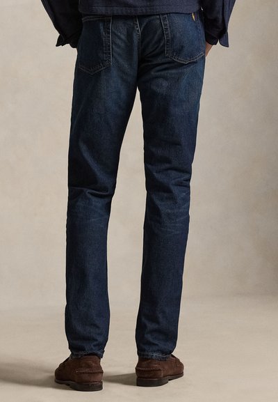 Asmuo, dėvintis tamsiai mėlynas džinsines kelnes ir rudus zomšinius batus, stovintis su rankomis kišenėse prieš neutralaus fono.