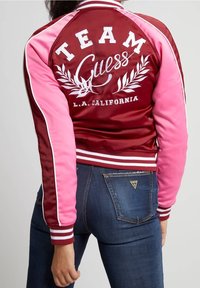 Rode en roze satijnen bomberjack met "TEAM Guess" borduursel, gestreepte manchetten en een aansluitend design. Gedragen met een donkere spijkerbroek.