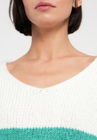 Pull en tricot avec un col en V, corps blanc et bande turquoise traversant la poitrine. Texture douce et coupe confortable.