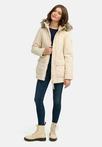Parka beige clair avec une capuche en fausse fourrure, fermeture éclair et pression, quatre poches avant et une coupe légèrement cintrée. Porté par-dessus une chemise bleu marine et un jeans.