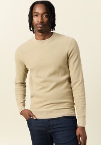 Maglione lavorato a coste beige con maniche lunghe e scollatura rotonda, abbinato a jeans blu scuro. Texture morbida, design aderente.