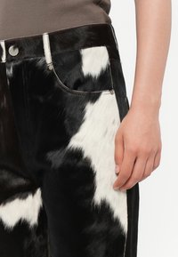 Jean imprimé vache noir et blanc avec une finition douce et texturée semblable à de la fourrure, comportant une poche avant et un détail de bouton métallique.