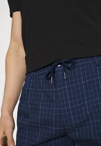 Námořnické modré kostičkované kalhoty s texturovanou tkaninou, vybavené stahovací šňůrkou v pase a stříbrnými akcenty. Na sobě je černé tričko.