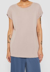 Ljusbeige, kortärmad topp i slätt tyg, med rund halsringning och upprullade ärmar. Bärs med lösa blå byxor.