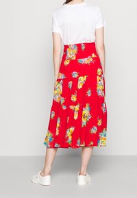 Roter Maxi-Rock mit gestuften Lagen, der eine Mischung aus bunten Blumenmustern zeigt; kombiniert mit einem weißen T-Shirt und weißen Sneakers.