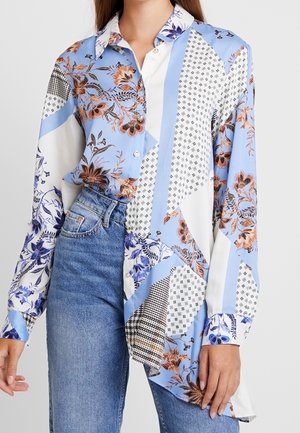 Chemise à manches longues à boutons avec des motifs floraux et géométriques mélangés en bleu, blanc et orange, portée rentrée dans un jean en denim bleu.
