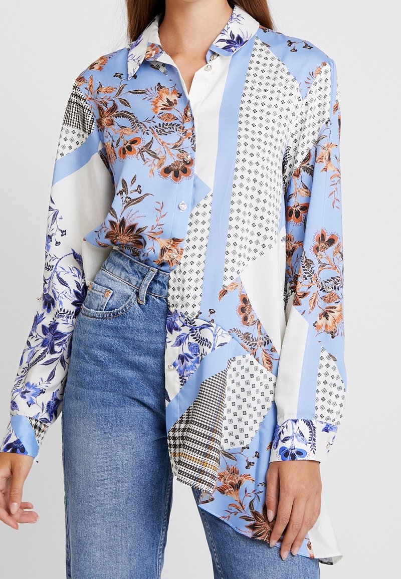 Chemise à manches longues à boutons avec des motifs floraux et géométriques mélangés en bleu, blanc et orange, portée rentrée dans un jean en denim bleu.