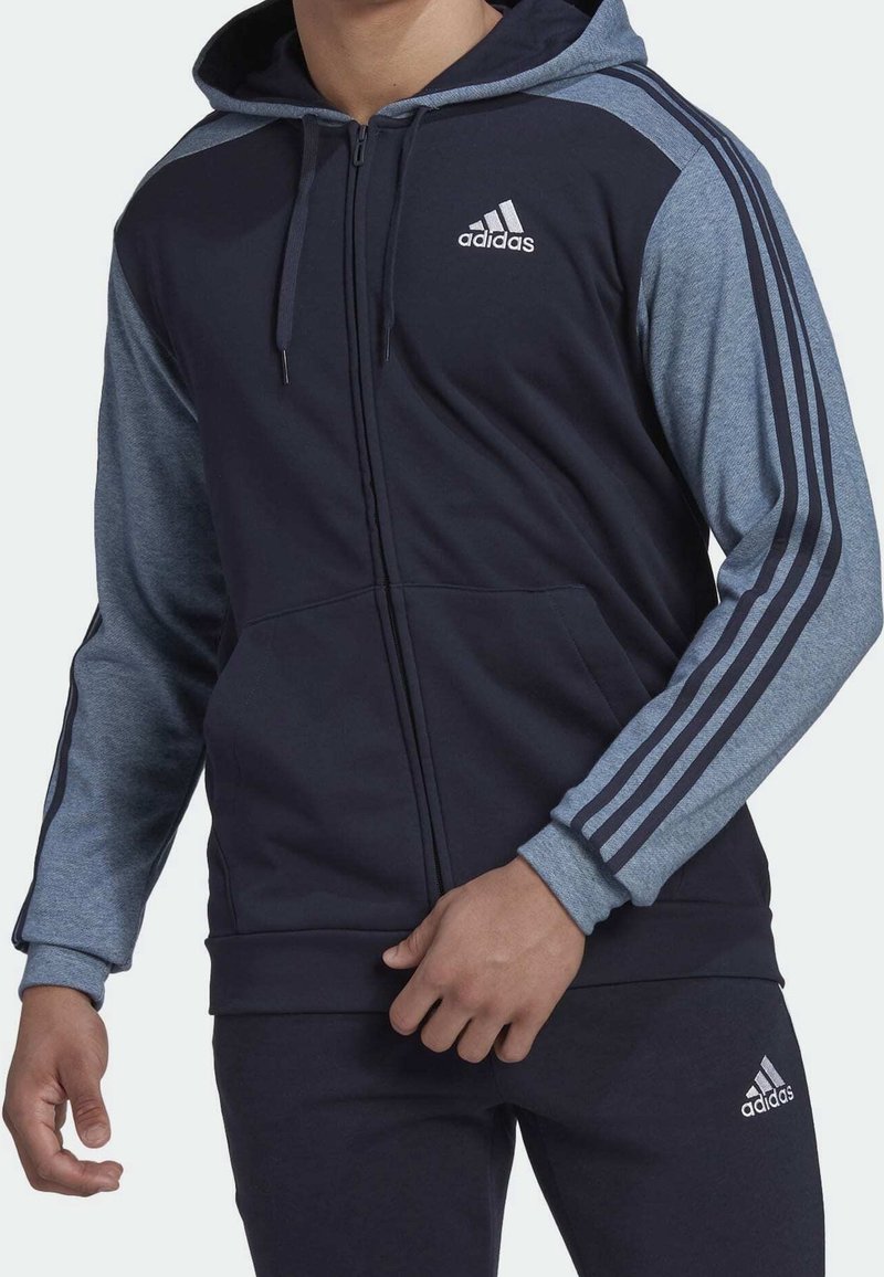 Homme portant un sweat à capuche zippé Adidas bleu marine et gris et un pantalon Adidas bleu marine assorti avec logo sur la poitrine et la cuisse.