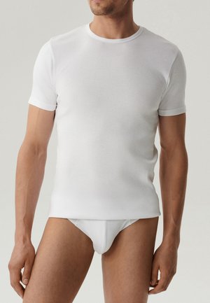 Mężczyzna ubrany w gładką białą koszulkę z krótkim rękawem i okrągłym dekoltem oraz białe majtki, stojący na jasnoszarym tle.