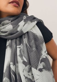 Harmaa ja valkoinen kuviollinen huivi resorikuvioisena, kiedottuna olkapään ympärille, jossa on abstrakteja kukkakuoseja ja pehmeää kangasta.