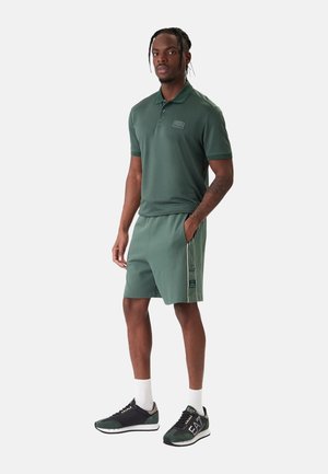 Uomo in piedi che indossa una polo verde scuro e pantaloncini abbinati con strisce laterali, abbinati a scarpe da ginnastica nere e verdi e calzini bianchi.