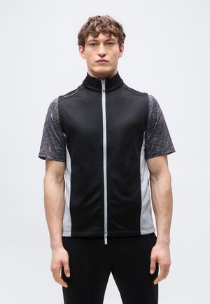 Ung mand står vendt fremad iført en sort og grå kortærmet vest med lynlås over en mønstret grå skjorte mod en ensfarvet baggrund.