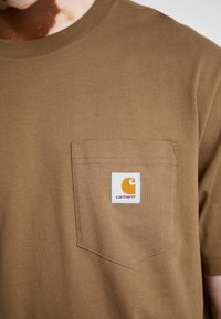 Barna pamut póló kerek nyakkal és bal oldali zsebbel. Az zseben kivágott Carhartt logó található narancssárga és fehér színben.