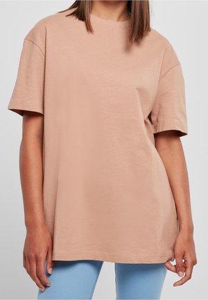 Kvinde iført løs, beige t-shirt med korte ærmer og lyseblå bukser, stående mod en neutral grå baggrund.