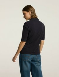 Granatowa koszulka polo z klasycznym kołnierzykiem, krótkimi rękawami i dopasowanym krojem, w zestawieniu z jasnoniebieskimi szerokimi dżinsami na jednolitym tle.