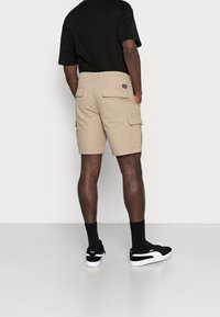 Homme portant un short cargo beige, un t-shirt noir, des chaussettes noires et des baskets Puma noires, se tenant contre un fond blanc, les mains dans les poches.