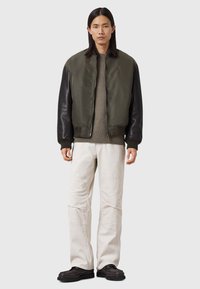 Homme portant une veste bomber olive zippée avec des manches noires, un pull brun, un pantalon blanc et des chaussures noires, se tenant contre un fond uni.