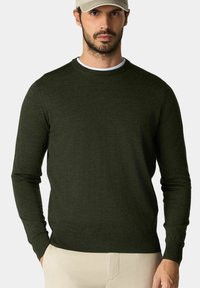 Tmavě zelený svetr s dlouhým rukávem z měkké pleteniny, s kulatým výstřihem, s ribkovanými manžetami a spodním lemem, v kombinaci se béžovými kalhotami.