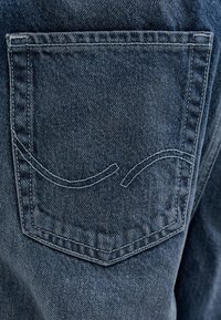 Poche arrière de jean en denim avec des coutures doubles et un motif de points en ligne courbe et brisée en fil bleu clair.