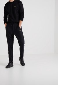 Svart sweatshirt med liten guldig logotyp, tillsammans med svarta joggerbyxor med sidofickor. Svarta sneakers kompletterar looken.