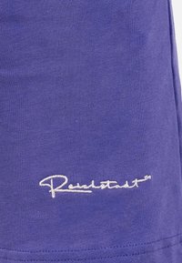 Tessuto viola con una consistenza liscia presenta un marchio ricamato bianco vicino all'orlo. Il design è semplice e privo di ornamenti.