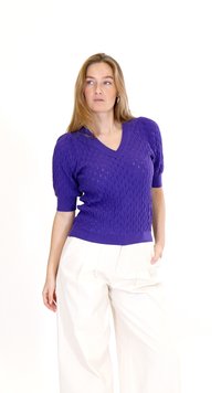 Sieviete ar violets teksturētu īsroku džemperi un vaļīgām baltām biksēm, stāv ar vienu roku kabatā, skatās uz sāniem.