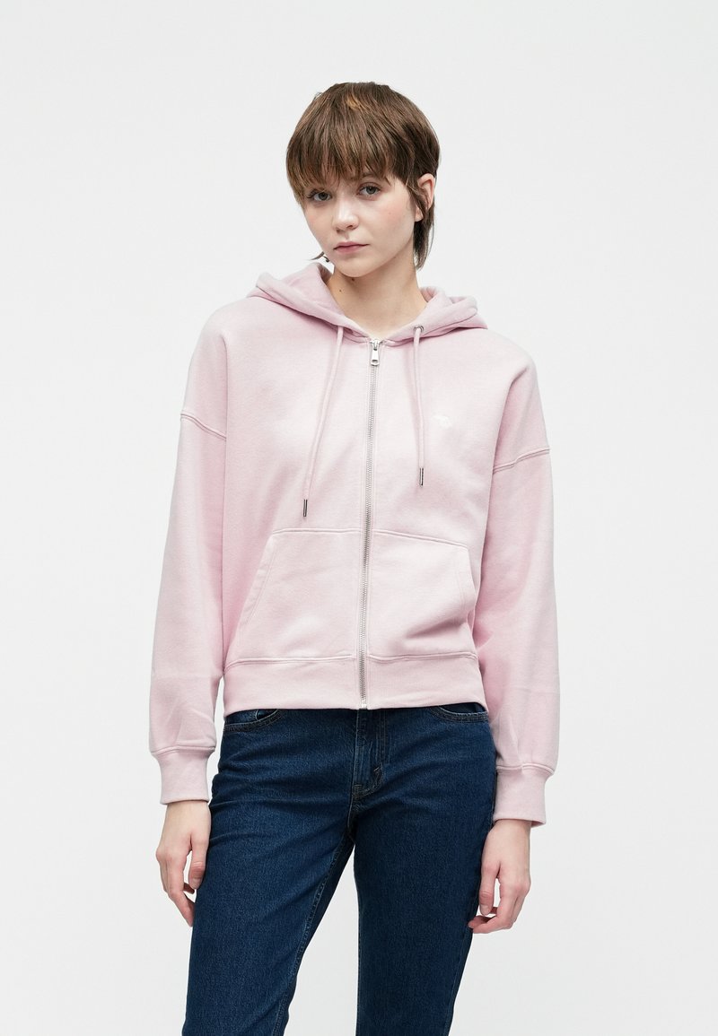 Young person wearing a light pink zip-up hoodie with front pockets and dark blue jeans, standing against a plain white background.