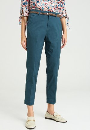 Femme portant un pantalon sarouel bleu sarcelle avec une ceinture tressée marron, un chemisier floral avec des manches nouées, et des mocassins crème avec un détail de chaîne en or.