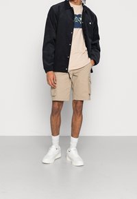 Tmavě hnědá kordrová košile/jacket, béžové tričko s potiskem, béžové cargo kraťasy, bílé tenisky a ponožky, stojící na neutrálním pozadí.