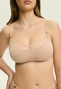 Beżowy bezprzewodowy biustonosz z przezroczystymi siateczkowymi panelami, regulowanymi ramiączkami i metalowymi detalami. Gładka faktura i minimalistyczny design.