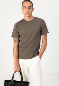 Braun kurzärmliges T-Shirt mit rundem Ausschnitt, kombiniert mit hellen Hosen. Eine schwarze Tote-Tasche wird in der linken Hand gehalten.