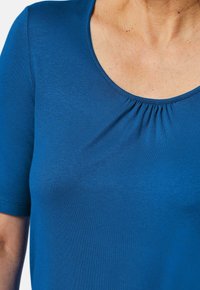 Blaues kurzärmliges Oberteil aus glattem Stoff, mit einem runden Halsausschnitt und Raffung in der Mitte.