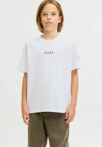 Valge puuvillane T-särk, millel on mereväe sinine "JACK & JONES" logo, lühikesed varrukad, ümar kaelus ja mugav lõige. Kombineeritud oliivroheliste pükstega.