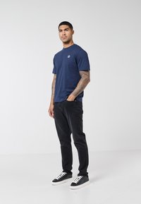 Man with tattooed arms wearing a navy blue t-shirt, black jeans, and black sneakers with white soles standing against a plain white background.