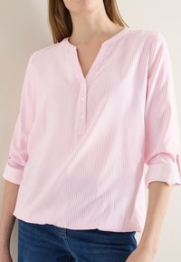 Femme portant un chemisier à rayures verticales rose clair et blanches avec trois boutons et manches retroussées, associé à un jean bleu foncé.