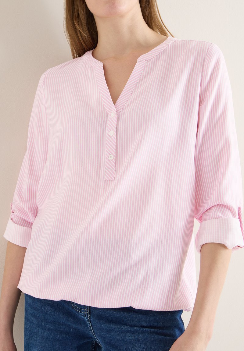 Femme portant un chemisier à rayures verticales rose clair et blanches avec trois boutons et manches retroussées, associé à un jean bleu foncé.