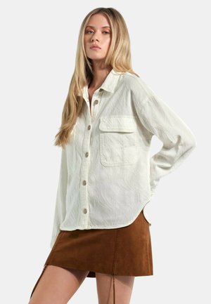 Giovane donna con lunghi capelli biondi che indossa una camicia bianca strutturata con bottoni e una mini gonna marrone in suede, posa con una mano sul fianco.