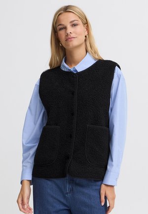 Femme blonde portant des boucles d’oreilles créoles en or, une chemise bleue à col, un gilet noir texturé sans manches boutonné et un jean bleu, debout devant un fond uni.